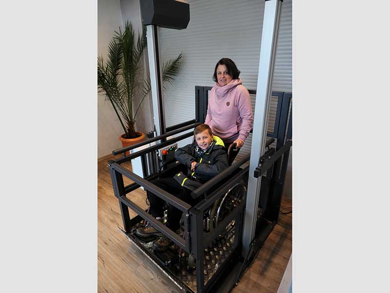 Plattformlift im Wohnraum mit einem Kind im Rollstuhl und einer Mutter dahinter